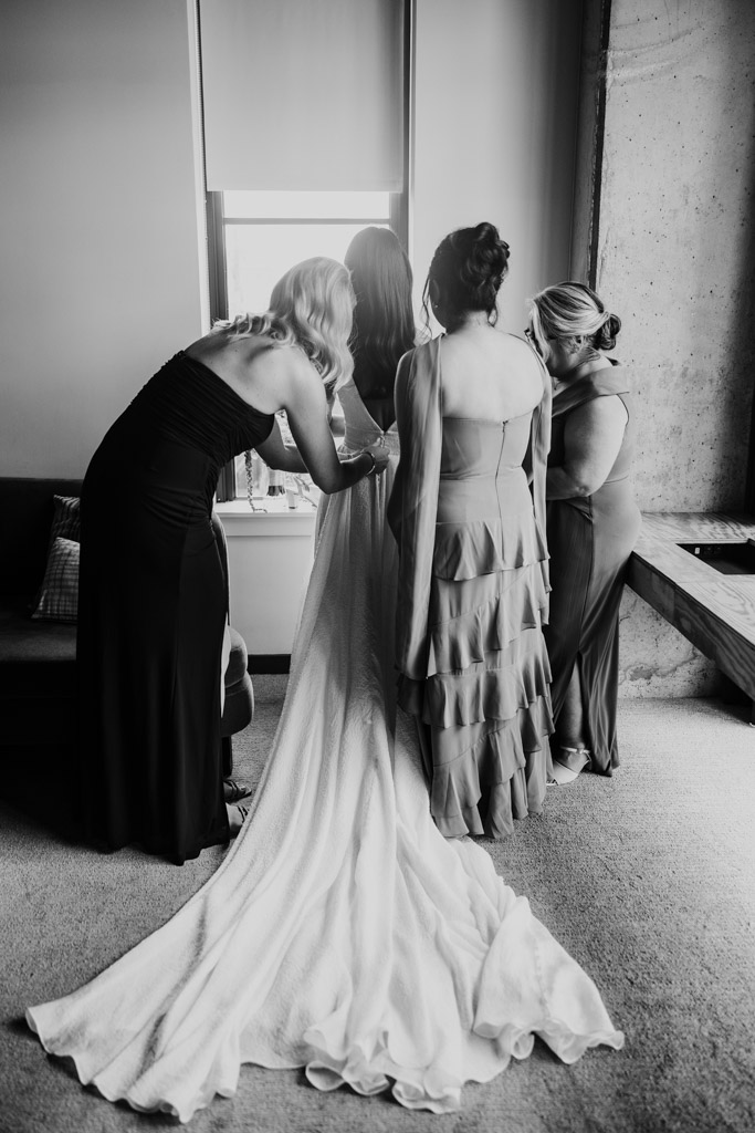Black and white photo of three women helping a bride in a long gown get ready by a window in a softly lit room before her Bottom Lounge wedding