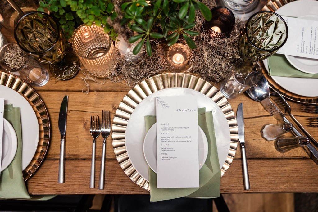 Guest place setting with gold-rimmed plates, green napkins, a menu card, and lush greenery centerpiece for Bottom Lounge wedding.reception