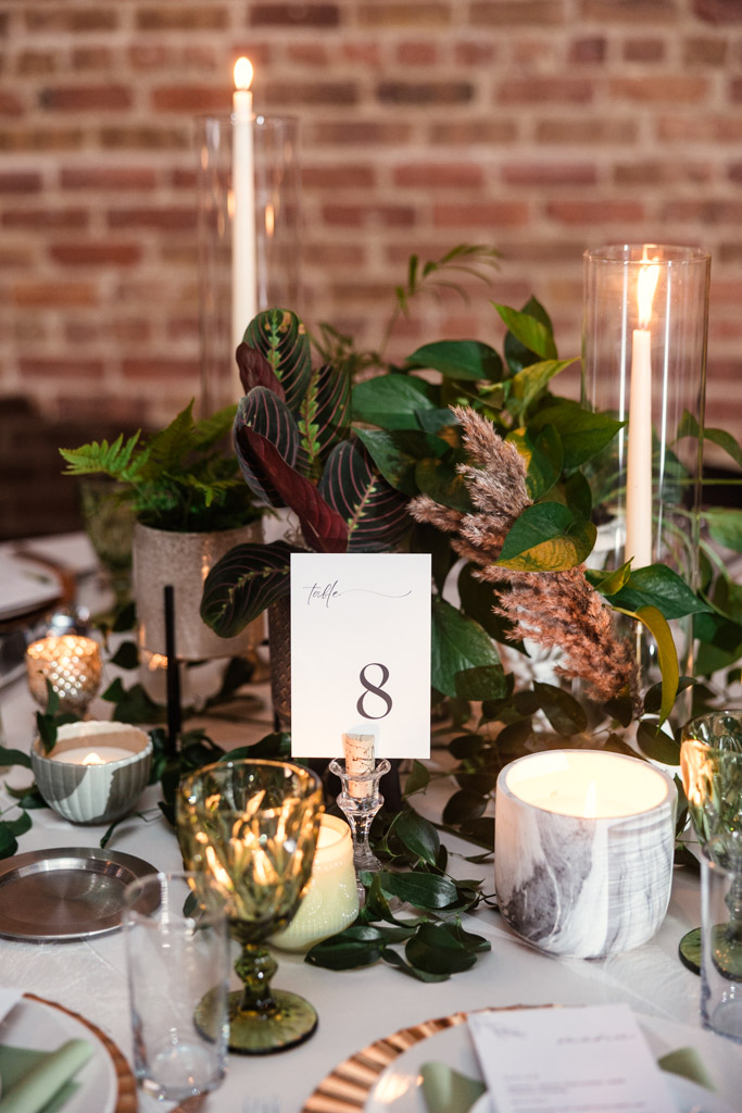 Table centerpiece with greenery, candles, and a table marker for a Bottom Lounge wedding reception against a brick wall background