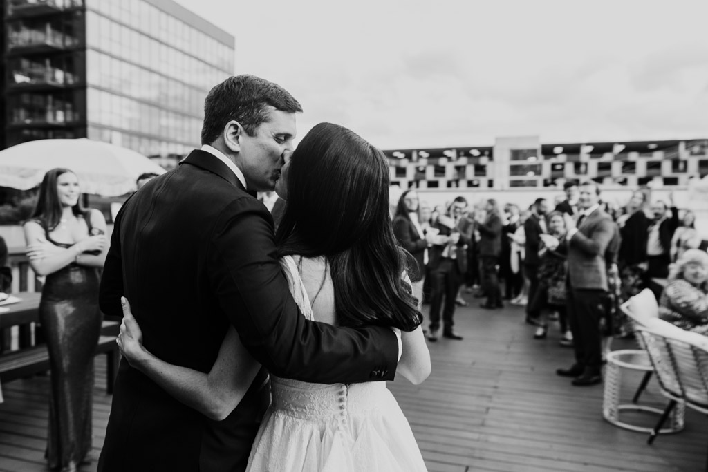 Black and white photo of newlywed couple embracing and kissing on a rooftop deck at their Bottom Lounge wedding, surrounded by a crowd of guests