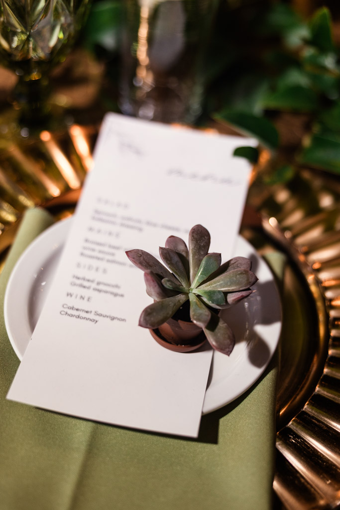 A dinner menu and a small succulent in a pot rest on a white plate with a gold charger underneath for Bottom Lounge wedding reception