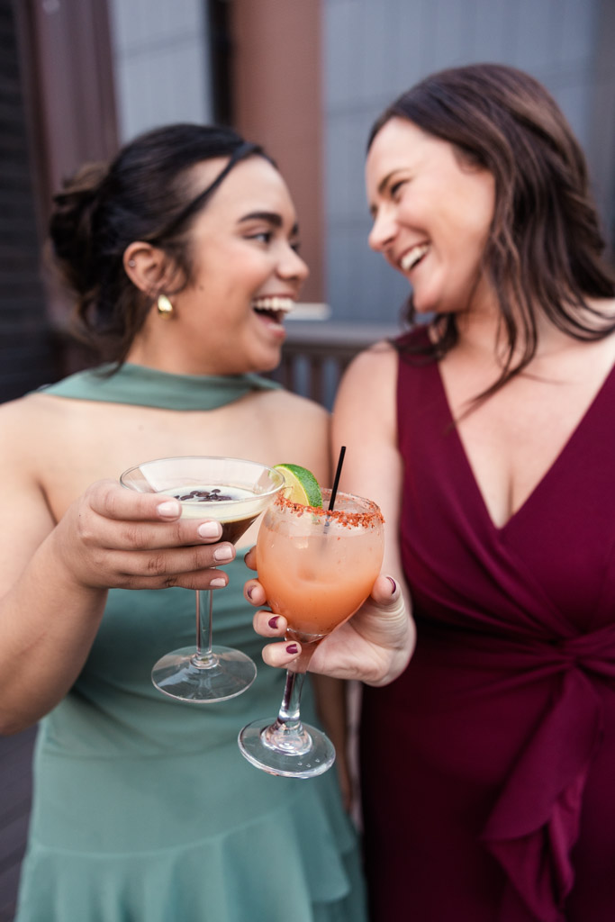 Two guests in green and burgundy dresses smiling and toasting with custom cocktails at a Bottom Lounge wedding reception