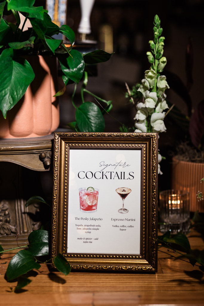 A framed custom cocktail menu on a table at a Bottom Lounge wedding reception surrounded by green plants and flowers