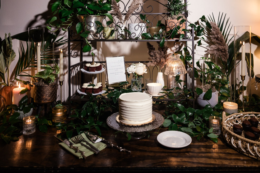 A white frosted wedding cake on a table with candles, greenery, cupcakes, and decorative plants for Bottom Lounge wedding reception