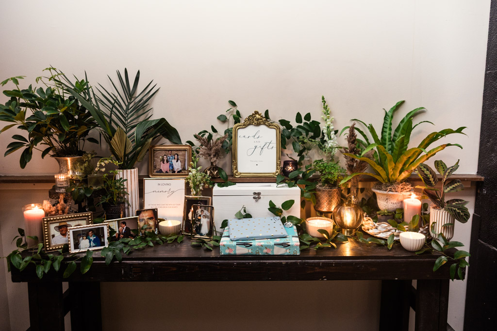 A decorated table with framed photos, plants, candles, and gift boxes arranged neatly against a white wall for Bottom Lounge wedding reception