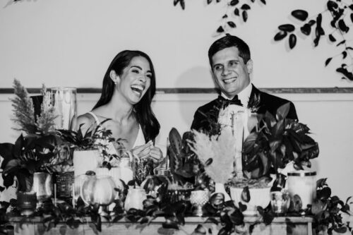 Black and white photo of bride and groom laughing behind a table decorated with candles and leafy plants at their Bottom Lounge wedding reception