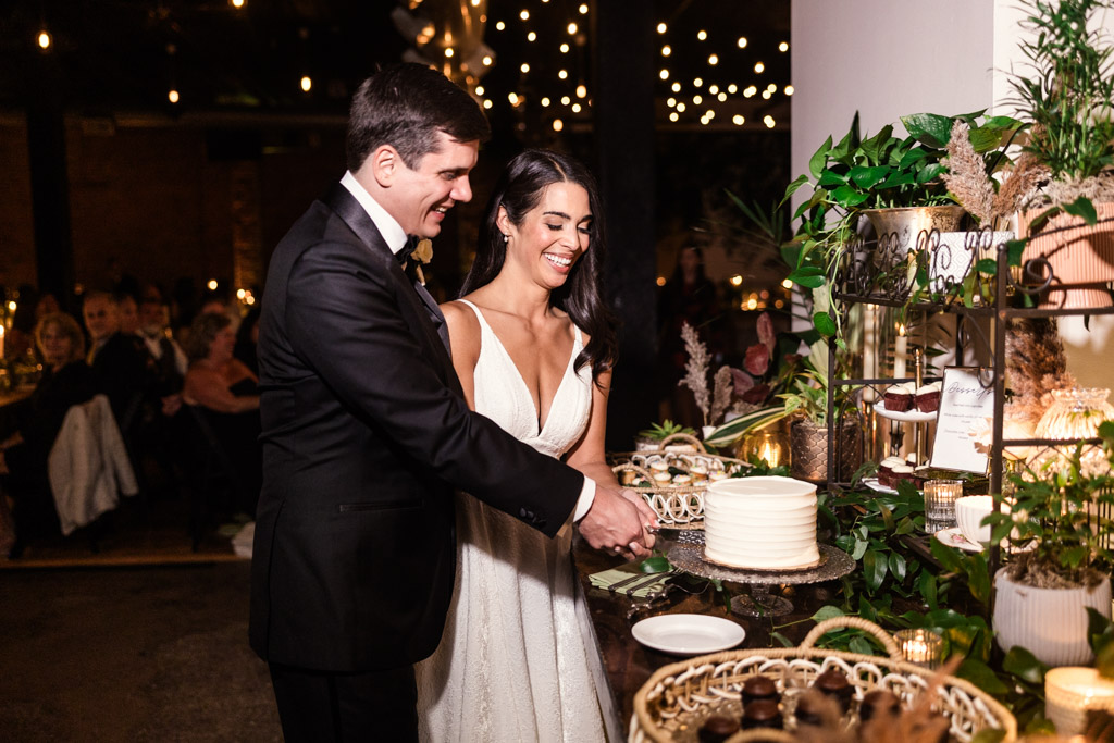 Bride and groom smiling as they cut their wedding cake together at a Bottom Lounge wedding reception with string lights