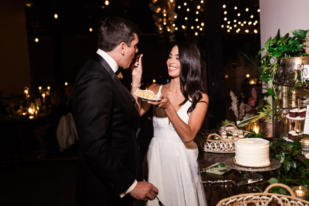 A bride feeds cake to the groom at their Bottom Lounge wedding reception, smiling under string lights