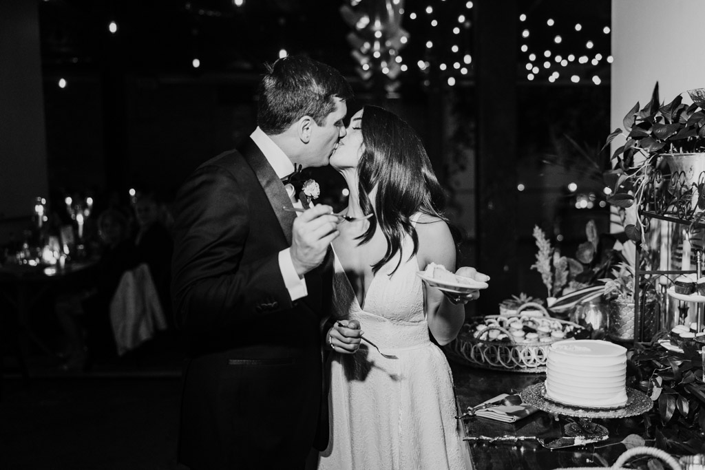 Black and white photo of newlywed couple in formal attire kissing while holding cake at their Bottom Lounge wedding reception