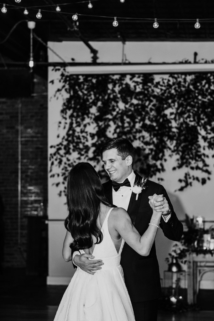 black and white photo of newlyweds during their first dance, smiling, under string lights at their Bottom Lounge wedding reception