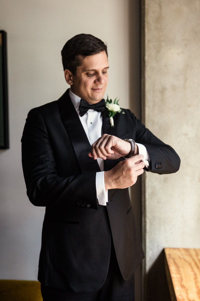 Groom in a black tuxedo adjusting his cuff links, standing indoors near a window with natural light, preparing for his Bottom Lounge wedding