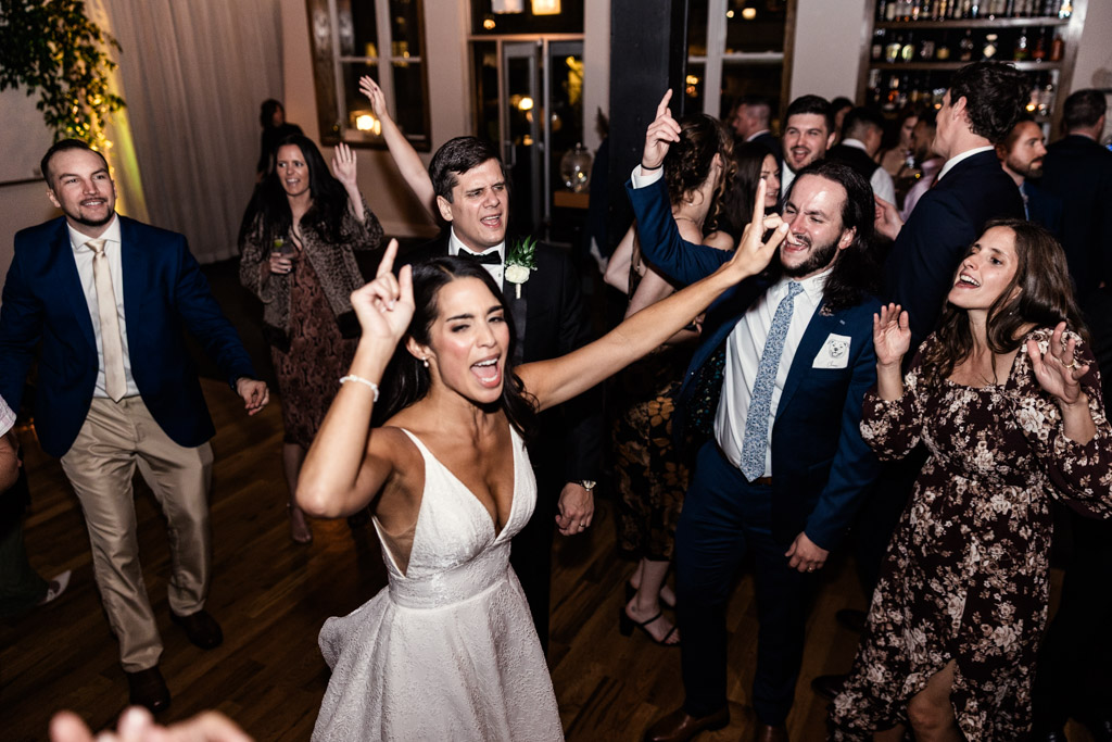 A bride and guests dance joyfully at a Bottom Lounge wedding reception, raising their hands and smiling.