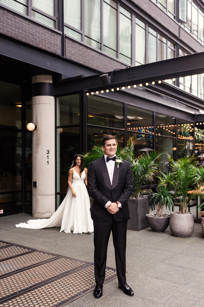 Groom in a tuxedo stands smiling as bride in a white gown approaches him during their first look outside The Emily Hotel