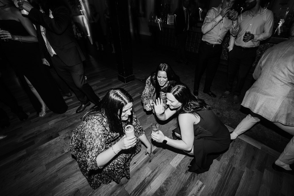 Black and white photo of three women crouch and laugh together at a Bottom Lounge wedding reception, holding drinks, while others dance nearby