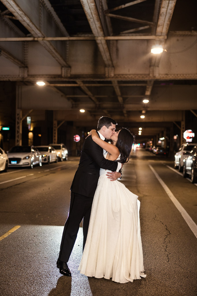Bride and groom kiss in the middle of a city street at night under an elevated train track, during their Bottom Lounge wedding reception