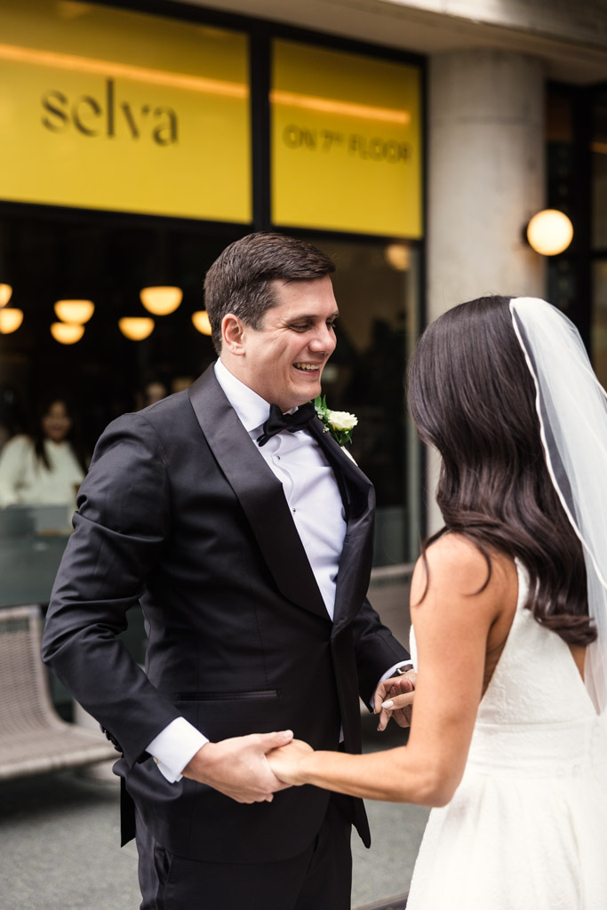 A groom smiles at his bride outdoors as they hold hands during their first look outside The Emily Hotel in Chicago