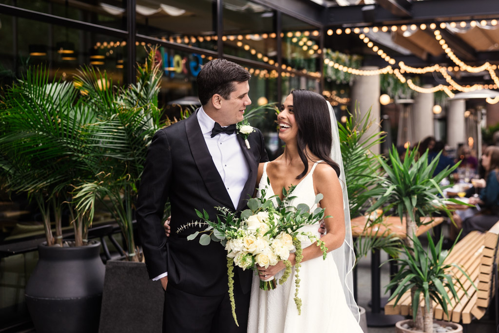 Bride and groom smiling at each other outside The Emily Hotel before their Bottom Lounge wedding, surrounded by green plants and glowing string lights