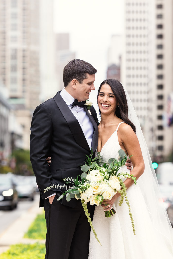 Bride and groom smiling, standing arm in arm on a Chicago street with tall buildings in the background