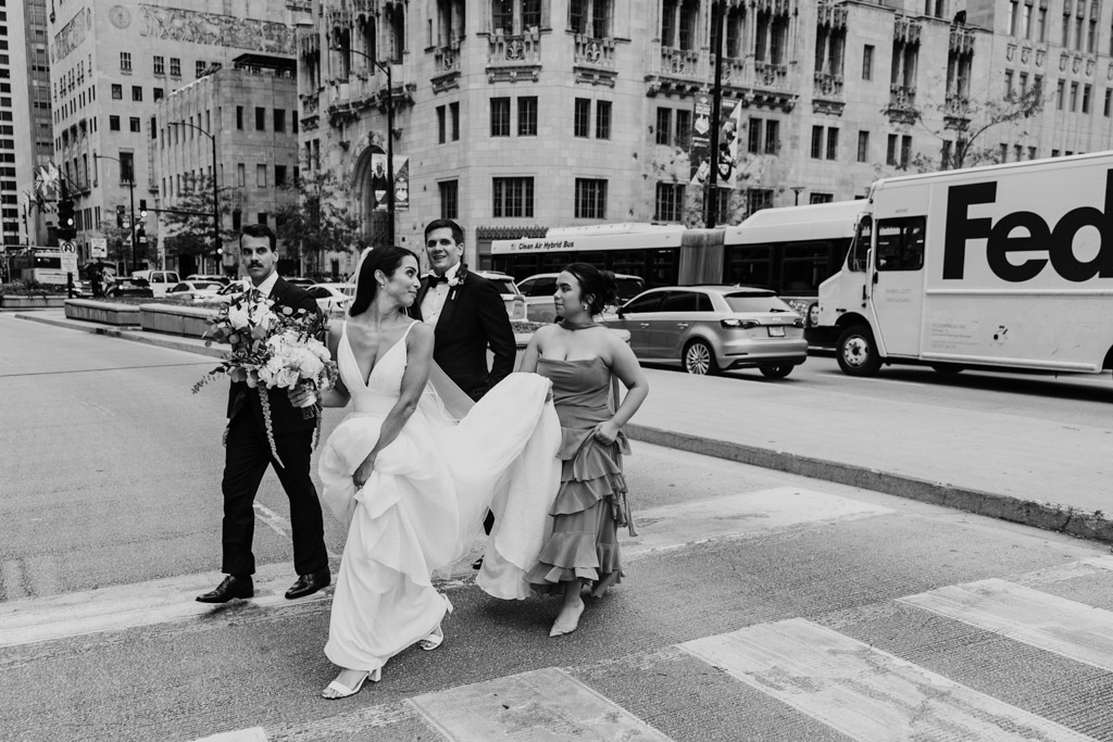 Black and white photo of bride, groom and wedding party crossing a downtown Chicago street, smiling and walking together with buildings and traffic behind them