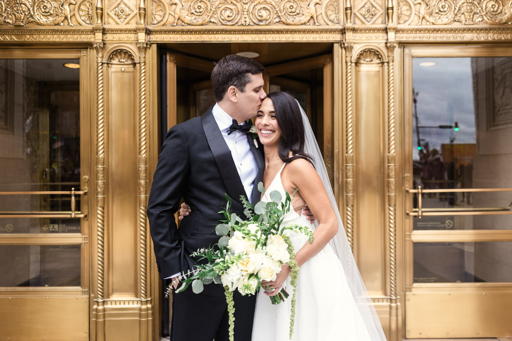 Groom kisses bride’s forehead as she smiles, holding a bouquet, in front of ornate gold doors at the Wrigley Building