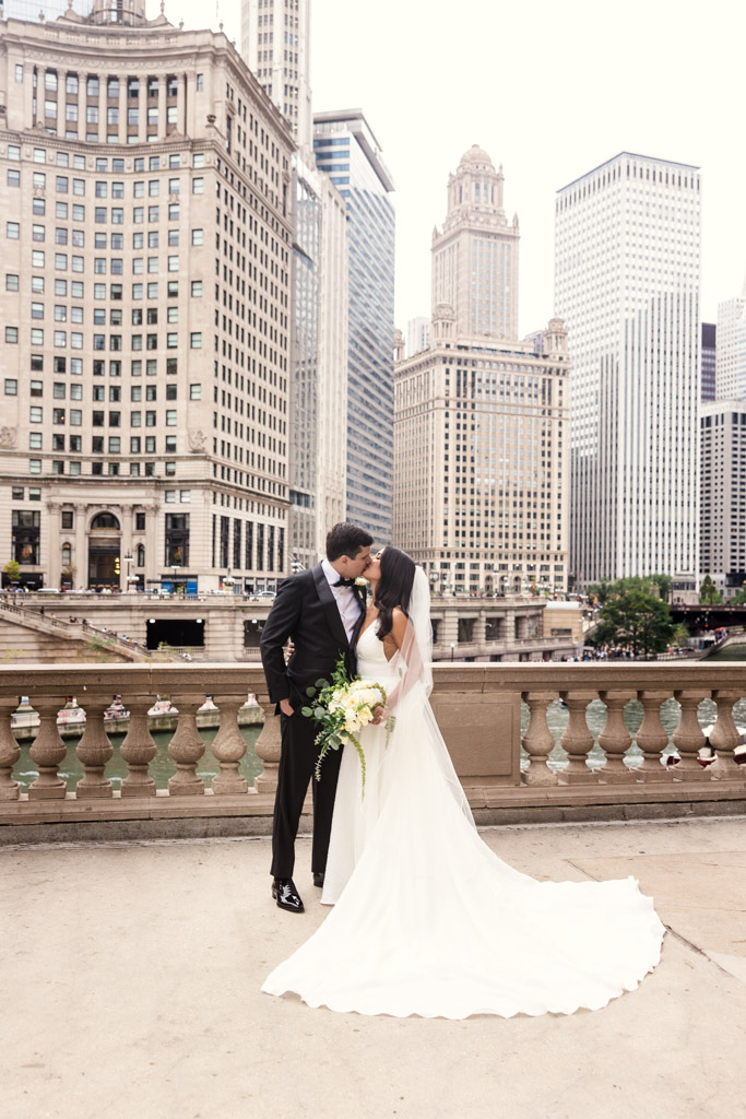 Bride and groom kiss outside the Wrigley Building near the Chicago River, tall buildings in the background