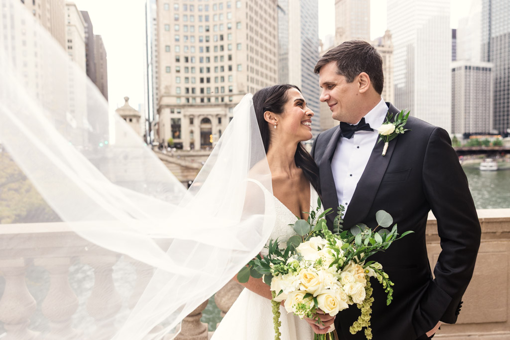 A bride and groom smile at each other while standing near the river in downtown Chicago, with bride's veil flowing in the wind