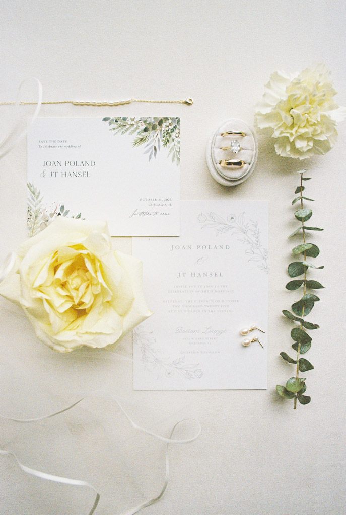 Flat lay of wedding invitations, yellow roses, rings, a carnation, earrings, and eucalyptus on a white background for Bottom Lounge wedding