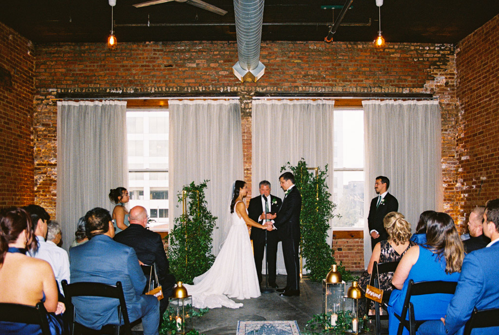 Film photo of bride and groom holding hands during an indoor Bottom Lounge wedding ceremony, surrounded by guests