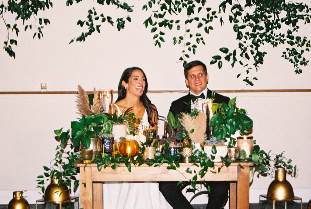 Film photo of happy newlyweds at a decorated table with green plants and gold accents, under hanging greenery, during their Bottom Lounge wedding reception