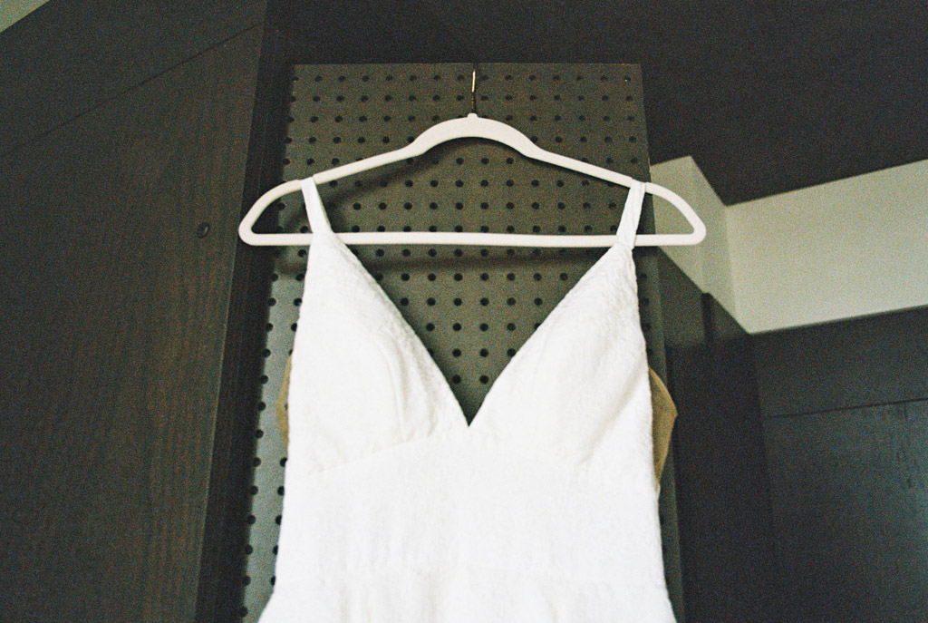 A sleeveless white dress with a V-neck hangs on a white hanger against a dark pegboard background for Bottom Lounge wedding
