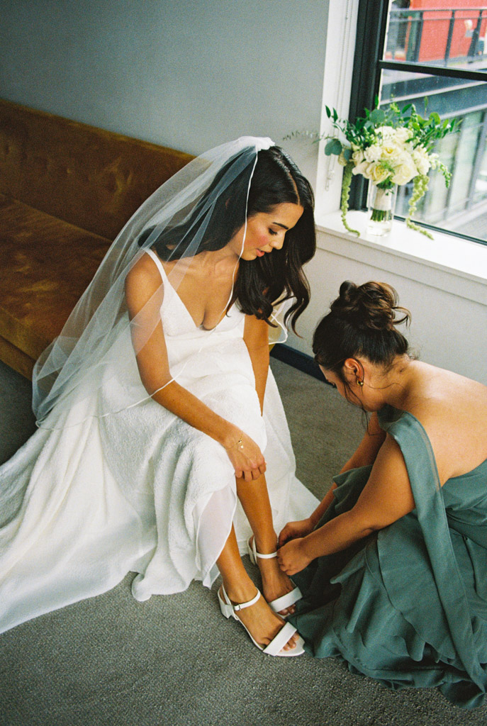 Film photo of bride in a white dress and veil sitting as a bridesmaid helps fasten her white shoe before her Bottom Lounge wedding