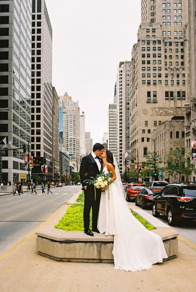 Film photo of bride and groom kissing on a city street median, surrounded by Chicago buildings and cars