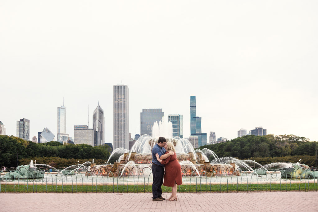 A couple embraces in front of a large fountain with a city skyline in the background, capturing their South Loop engagement.