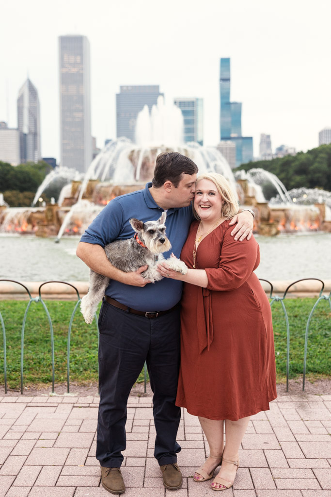 Engaged couple poses by Buckingham Fountain with their dog, Chicago skyline in the background