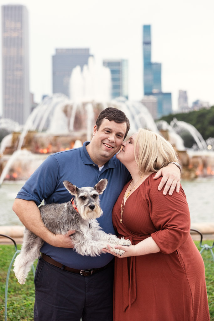 A woman kisses her partner’s cheek as he holds their dog with Buckingham Fountain in the background