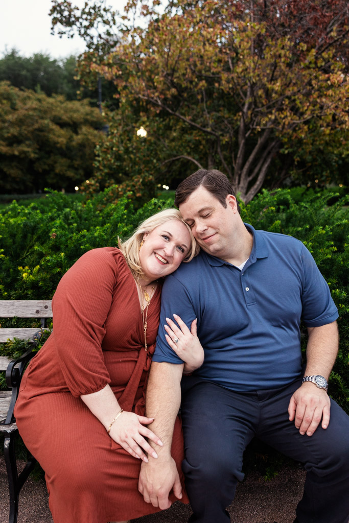 A smiling woman leans on a man's shoulder as they sit on a bench in Grant Park with green bushes and trees, during their South Loop engagement session