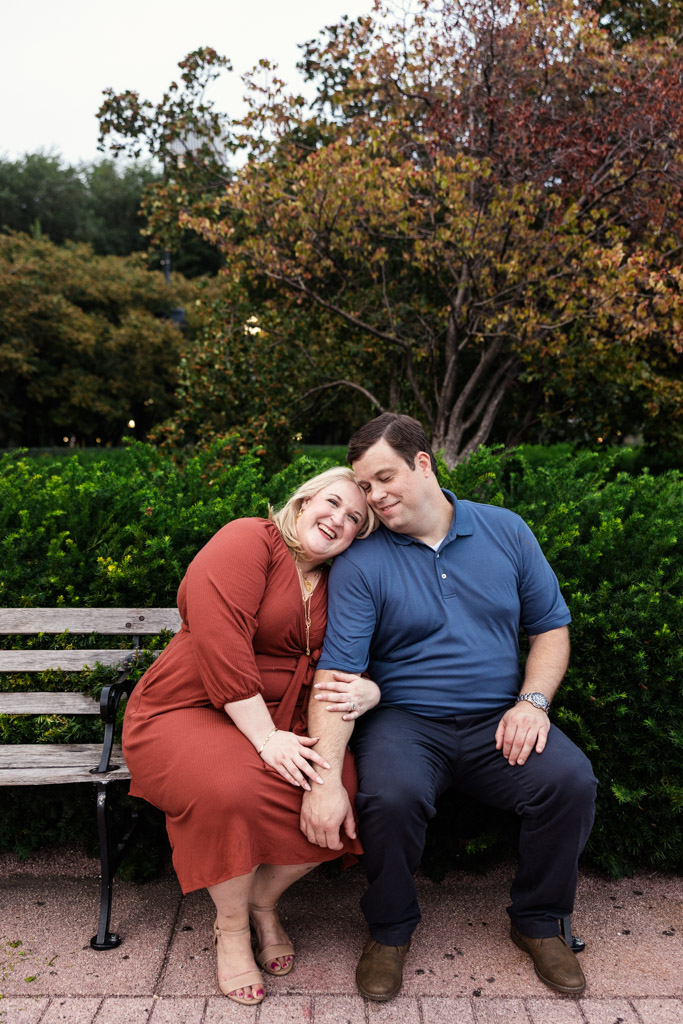 Happy engaged couple sits closely together on a Grant Park bench surrounded by green bushes and autumn trees.