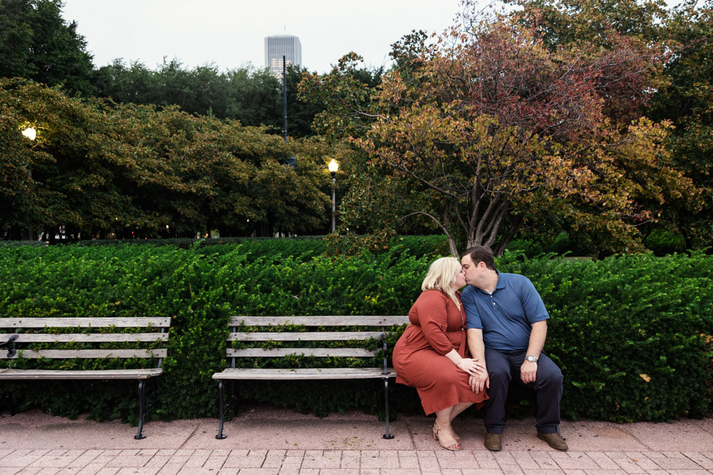 Engaged couple kisses on a park bench in Grant Park, surrounded by lush greenery and vibrant autumn trees