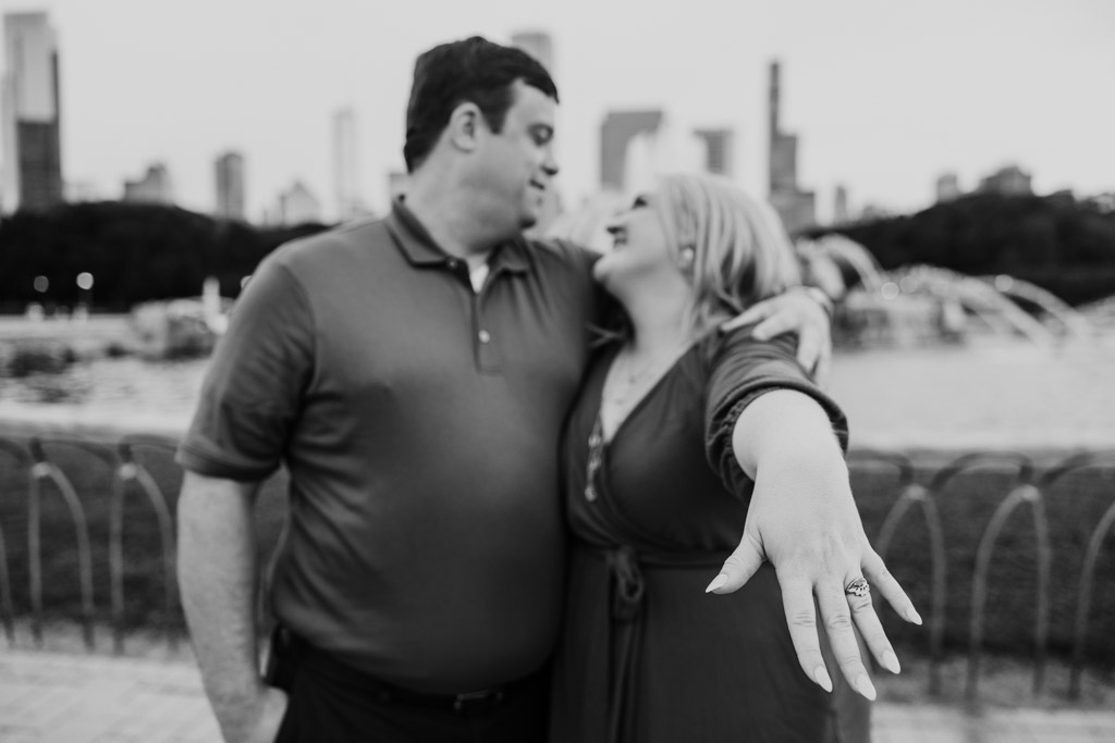 Black and white photo of woman smiling and showing off her engagement ring near Buckingham Fountain in Grant Park