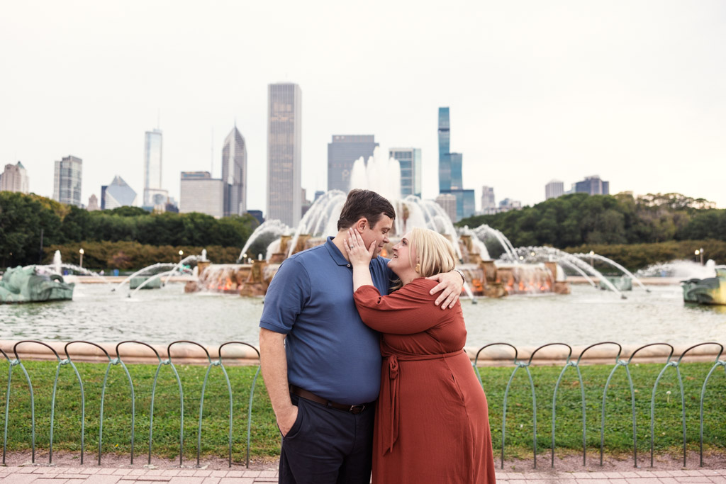 A couple embraces and smiles in front of Buckingham Fountain with Chicago skyline in the background during their South Loop engagement session