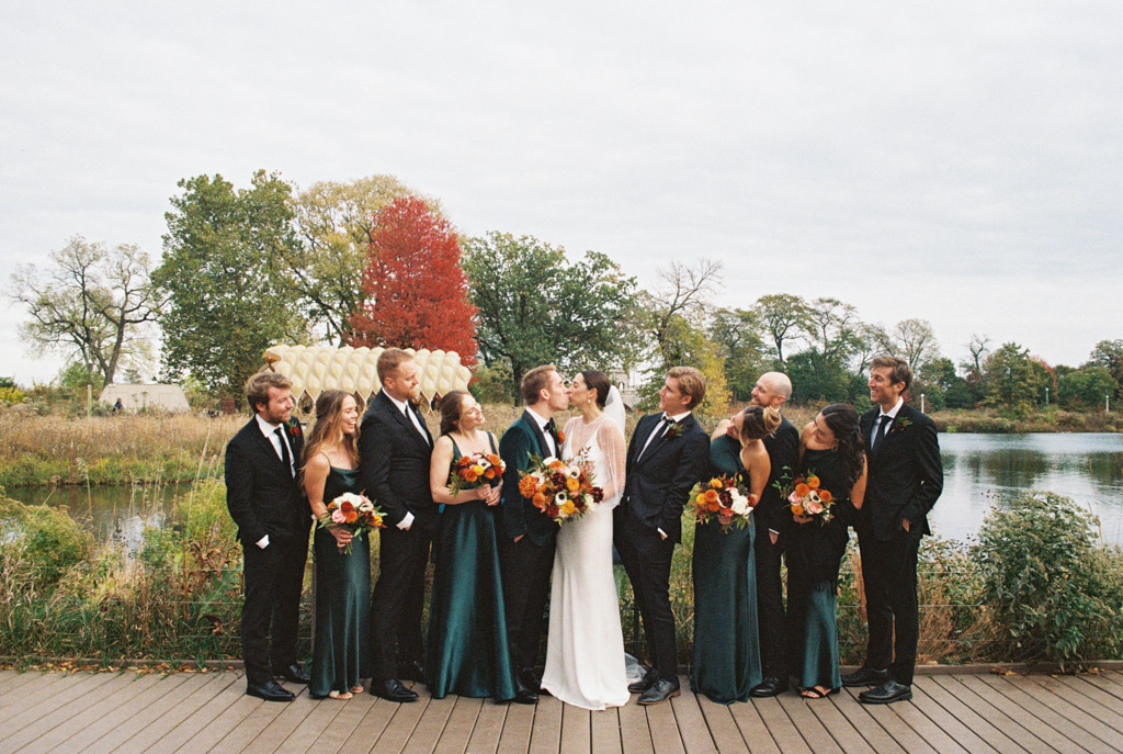Film photo of wedding party in formal attire on a deck by a Lincoln Park pond with the bride and groom kissing in the center