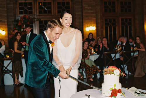 A smiling couple cuts a wedding cake together at their Cafe Brauer wedding reception, surrounded by applauding guests.
