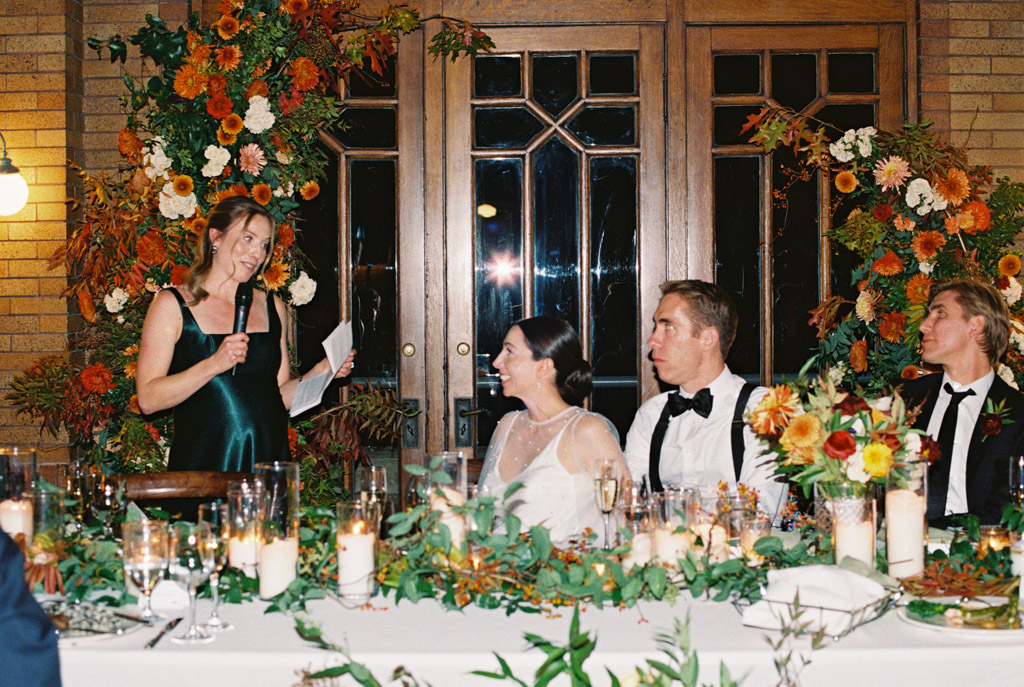 Film photo of Maid of Honor woman giving a speech to a smiling bride and groom at a flower-decorated table during their Cafe Brauer wedding reception