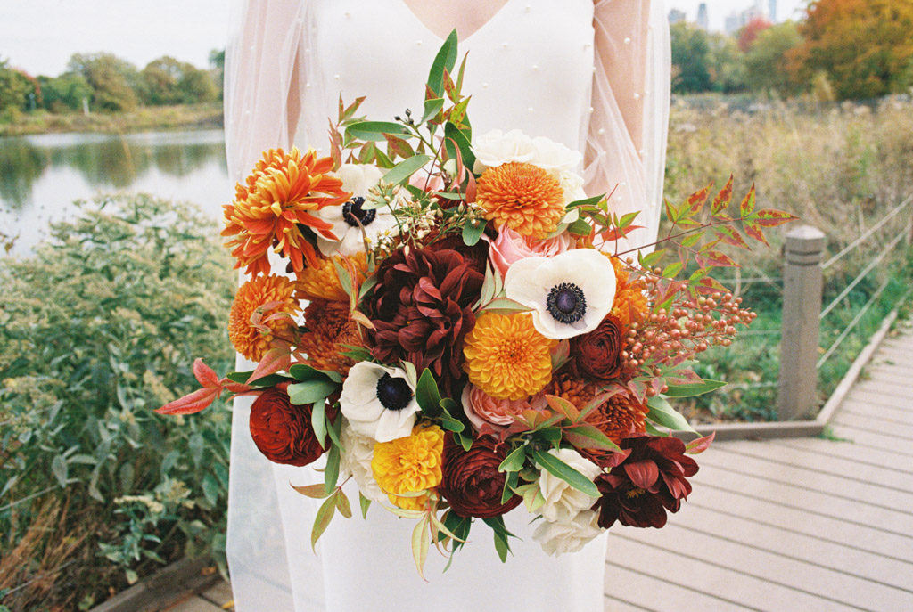 Film photo of bride holding a vibrant autumn bouquet with orange, yellow, white, and burgundy flowers on a boardwalk before her Cafe Brauer wedding celebration