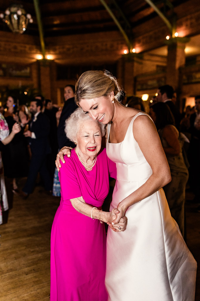Bride in white dress hugs and dances with older woman in bright pink dress at Cafe Brauer wedding reception