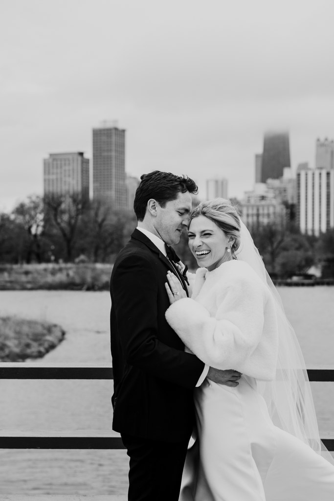Black and white photo of newlywed couple in wedding attire embracing and smiling in Lincoln Park with Chicago skyline in the background