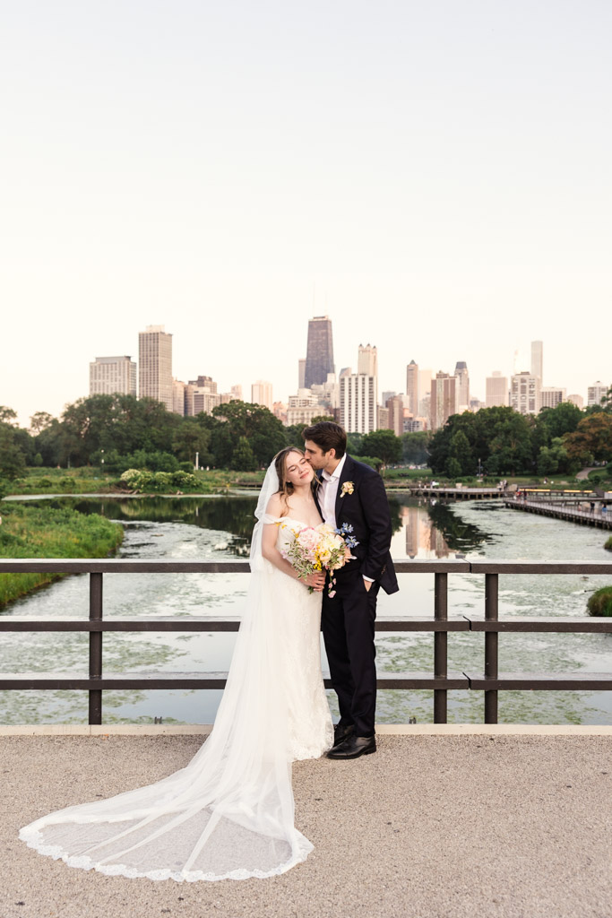 Bride and groom embrace on a Lincoln Park bridge with a city skyline and pond in the background