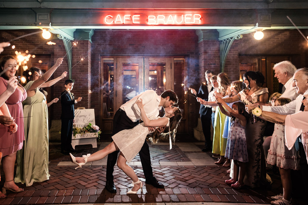 Newlywed couple kisses as they dance surrounded by cheering guests holding sparklers outside Cafe Brauer at night