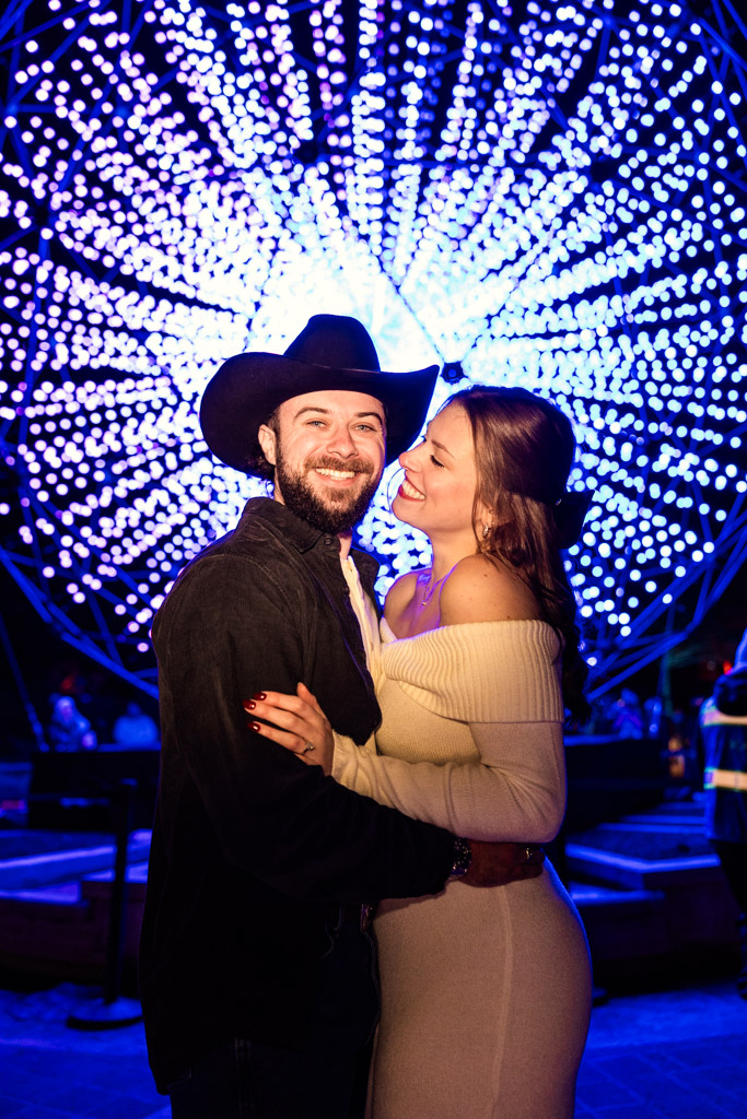 Engaged couple smiles and embraces in front of a large, glowing circular light display at night during Lightscape at Chicago Botanic Garden