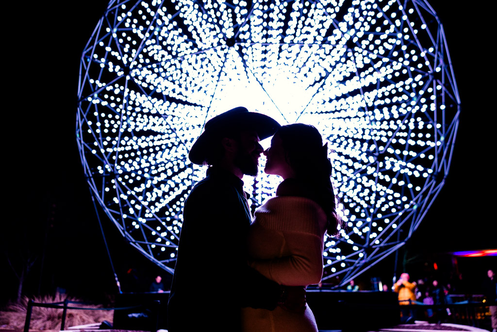 Engaged couple embraces in silhouette before a large, glowing spherical light display at night at Lightscape at Chicago Botanic Garden